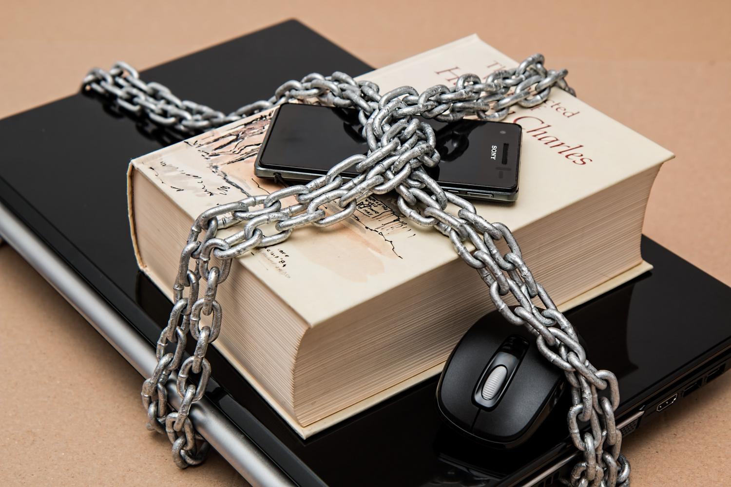 Cadena metálica envolviendo un portátil, un smartphone y un libro, como metáfora visual del cifrado y la seguridad digital.