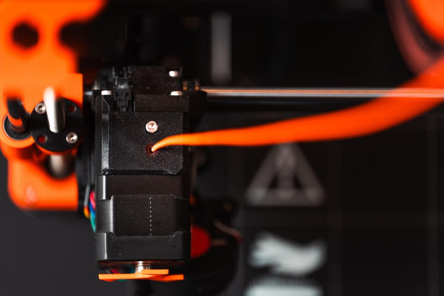 Primer plano detallado del extrusor de una impresora 3D con filamento naranja, mostrando el mecanismo de empuje del material.