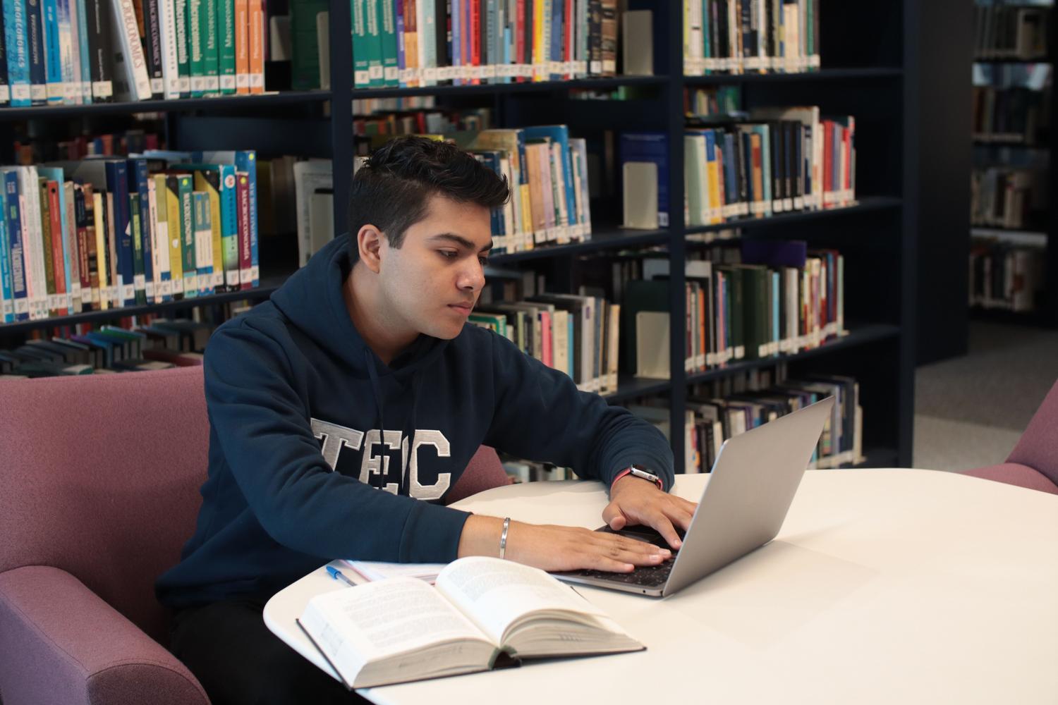Estudiante utilizando un ordenador portátil y libros en una biblioteca, representando el apoyo académico y profesional.