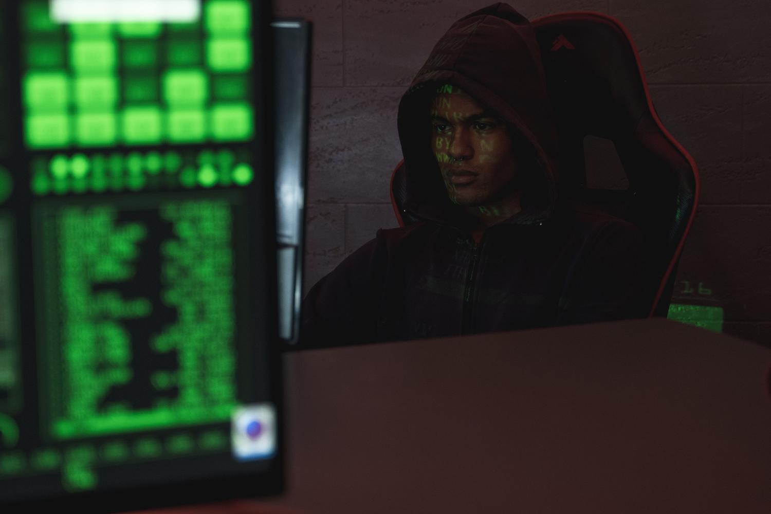 Programador con capucha concentrado frente a un monitor, representando la actividad de hackers y la ciberinvestigación.