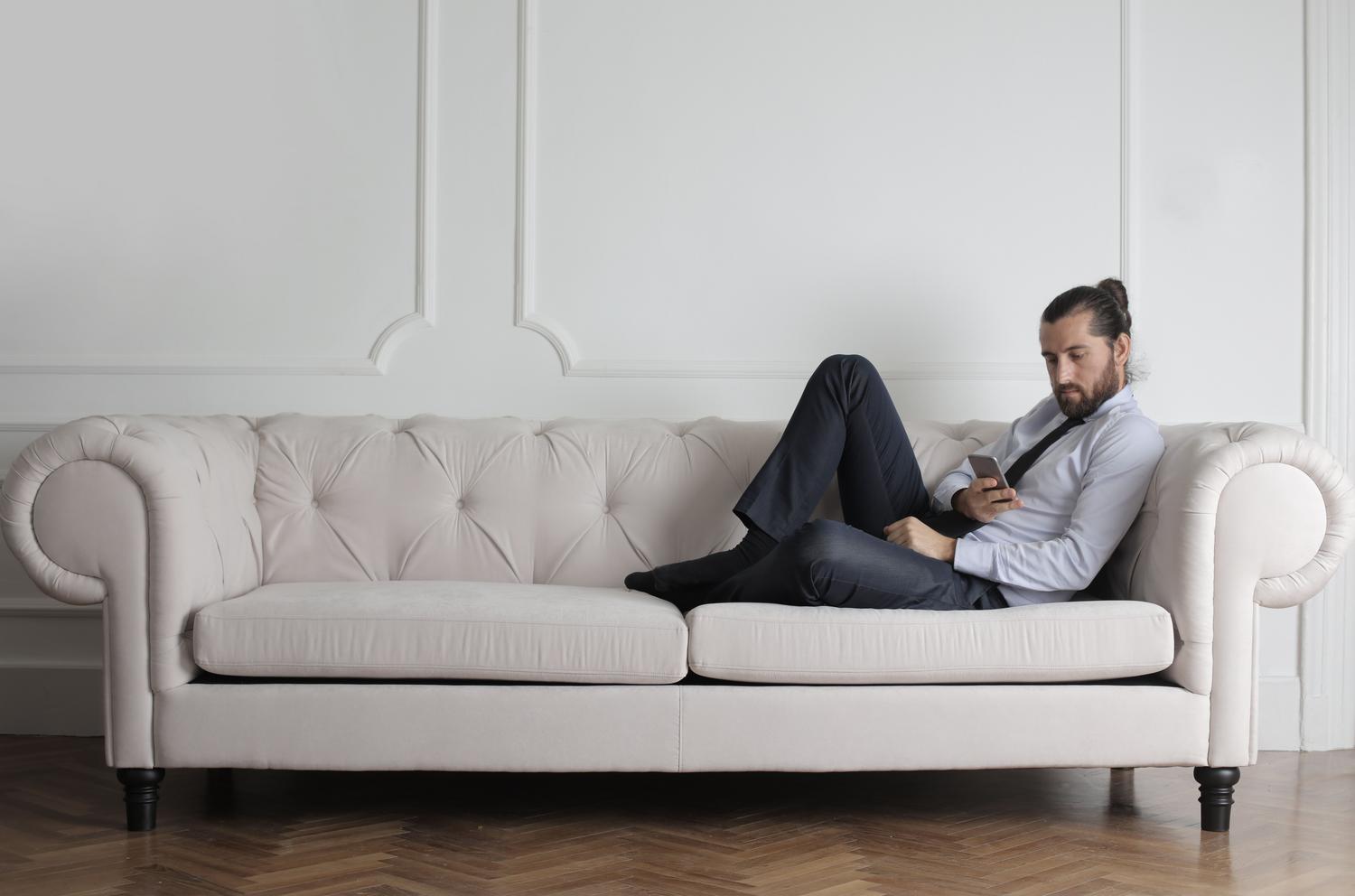 Hombre relajado descansando en un sofá blanco moderno mientras usa su teléfono móvil tranquilamente.