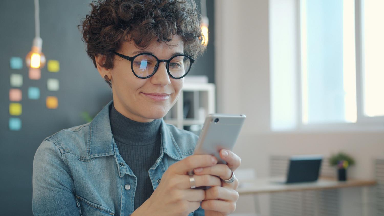 Mujer con gafas utilizando un smartphone, ilustrando la importancia de la salud visual y el uso de lentes protectoras contra la luz azul.