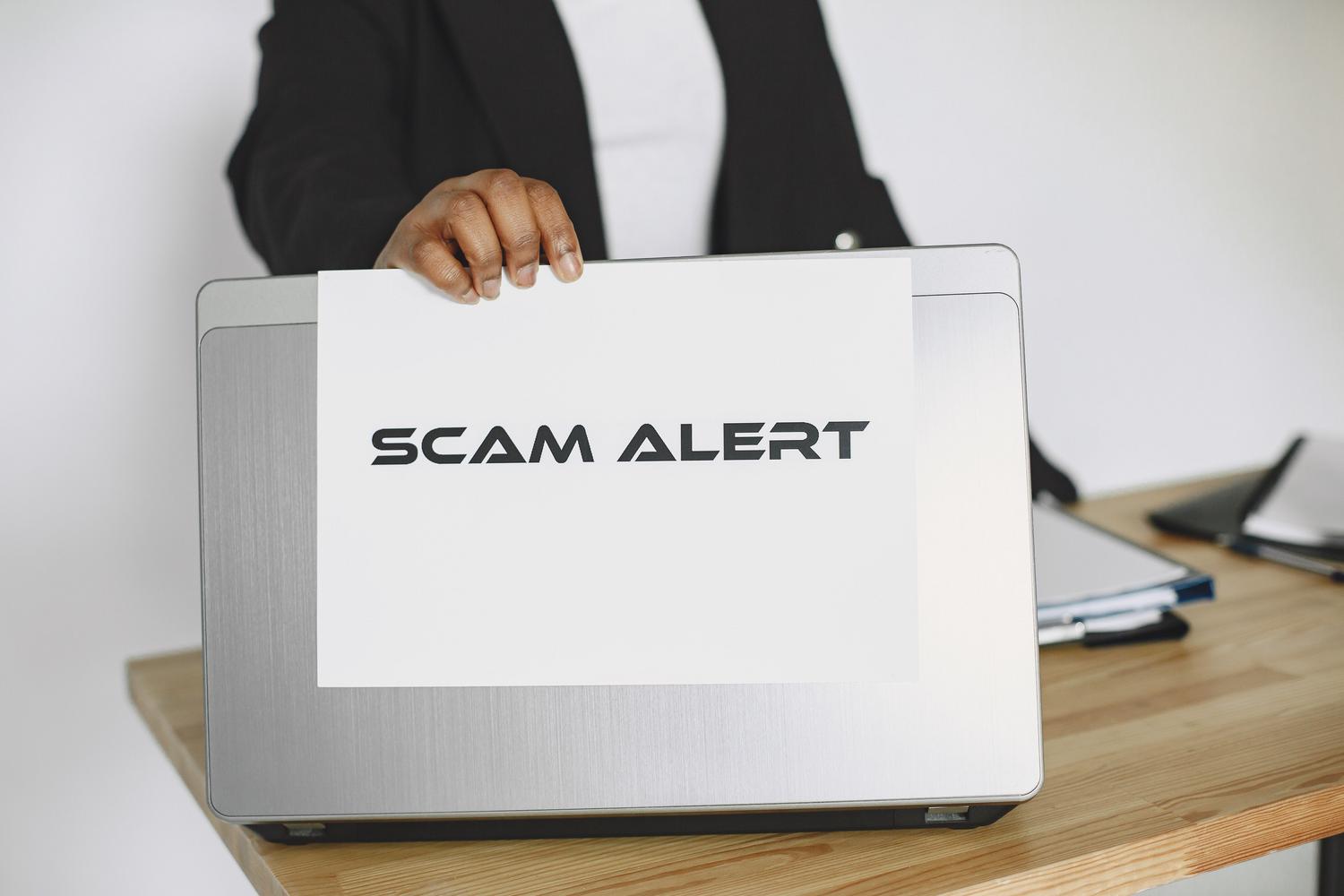 Una persona tiene in mano un cartello con la scritta "Attenzione truffa" sopra un computer portatile, avvertendo sui rischi di frode online e CAPTCHA falsi.