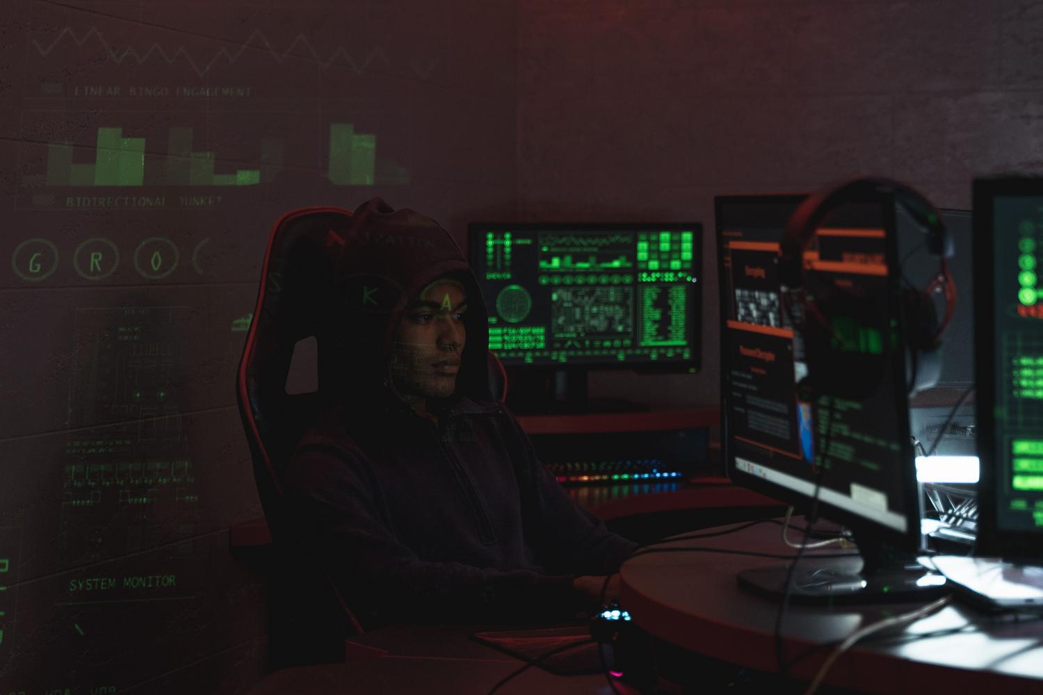 Hacker anónimo con capucha frente a múltiples pantallas con código, ilustrando el riesgo de filtraciones de datos y la Dark Web.
