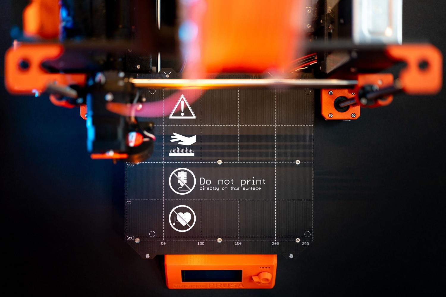 Detalle de la base de impresión de una impresora 3D con señalización de seguridad, ilustrando los costes operativos y de mantenimiento. Detalle de la base de impresión de una impresora 3D con señalización de seguridad, ilustrando los costes operativos y de mantenimiento.