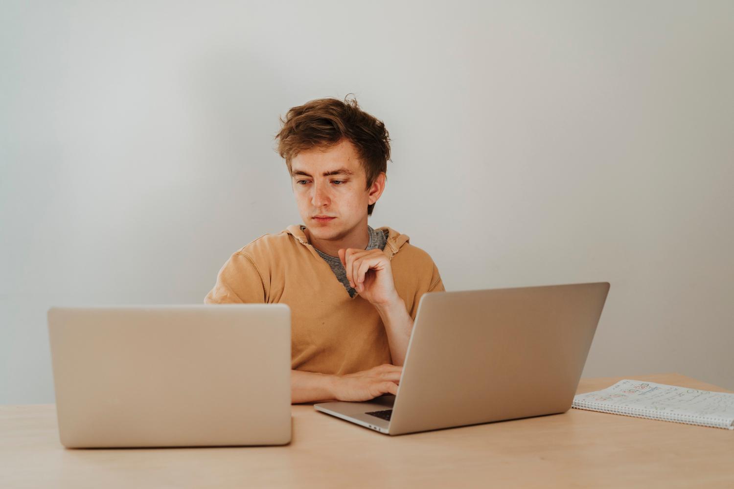 Joven estudiante universitario concentrado trabajando con dos portátiles y un cuaderno de notas