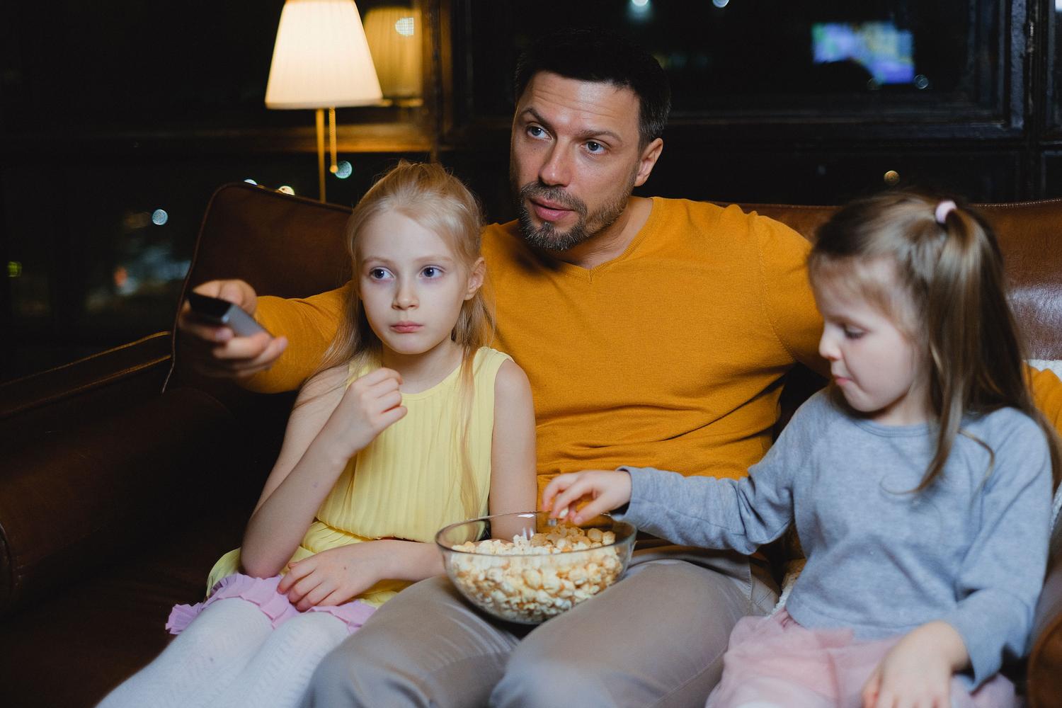 Padre e hijas disfrutando de una noche de cine con palomitas en el sofá, ilustrando la experiencia de consumo de contenido multimedia en familia.