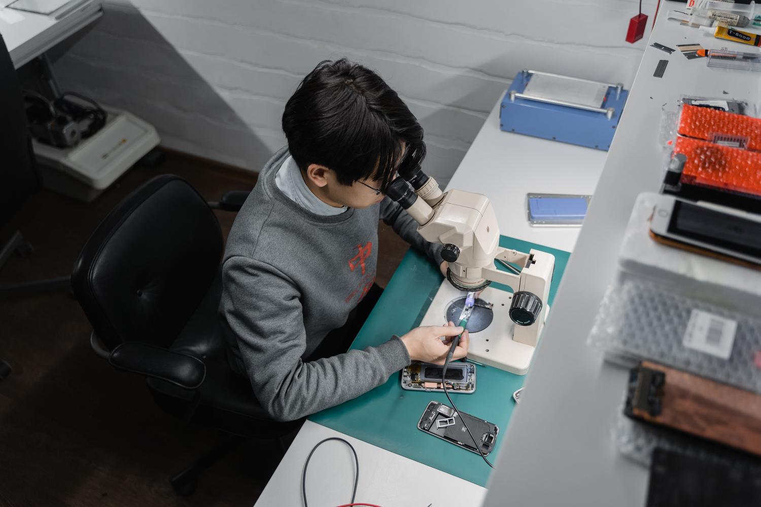 Técnico profesional reparando un dispositivo móvil con herramientas de precisión y microscopio.