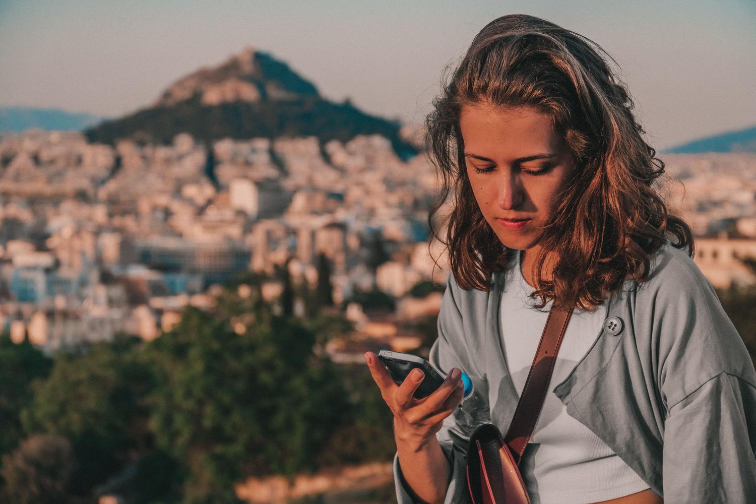 Una turista utilizando su teléfono móvil con la ciudad de Atenas de fondo, representando el uso de aplicaciones de traducción en el extranjero.