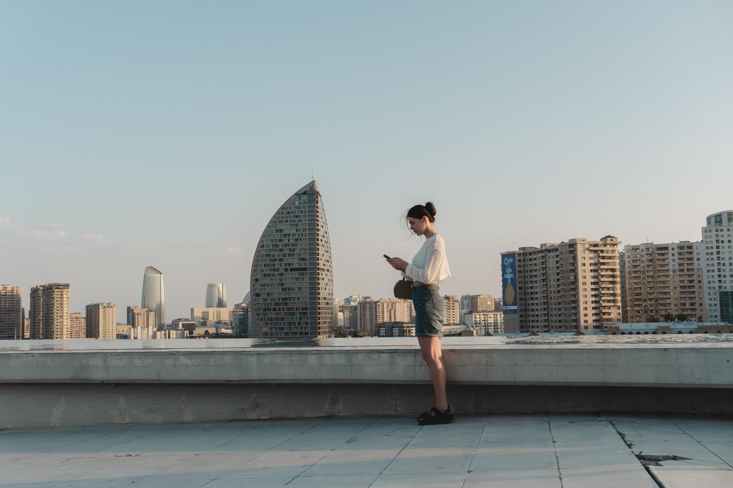Mujer joven revisando su smartphone frente a un moderno skyline urbano, ilustrando el uso de tecnología móvil durante el turismo.