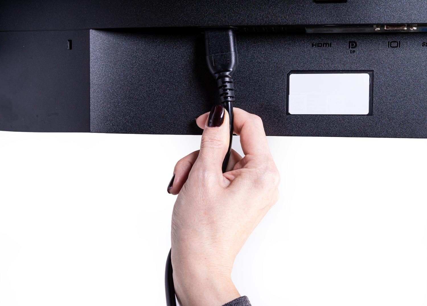 Imagen de una mano conectando un cable en la parte trasera de un monitor, donde se aprecian las etiquetas de los puertos HDMI y DisplayPort (DP).