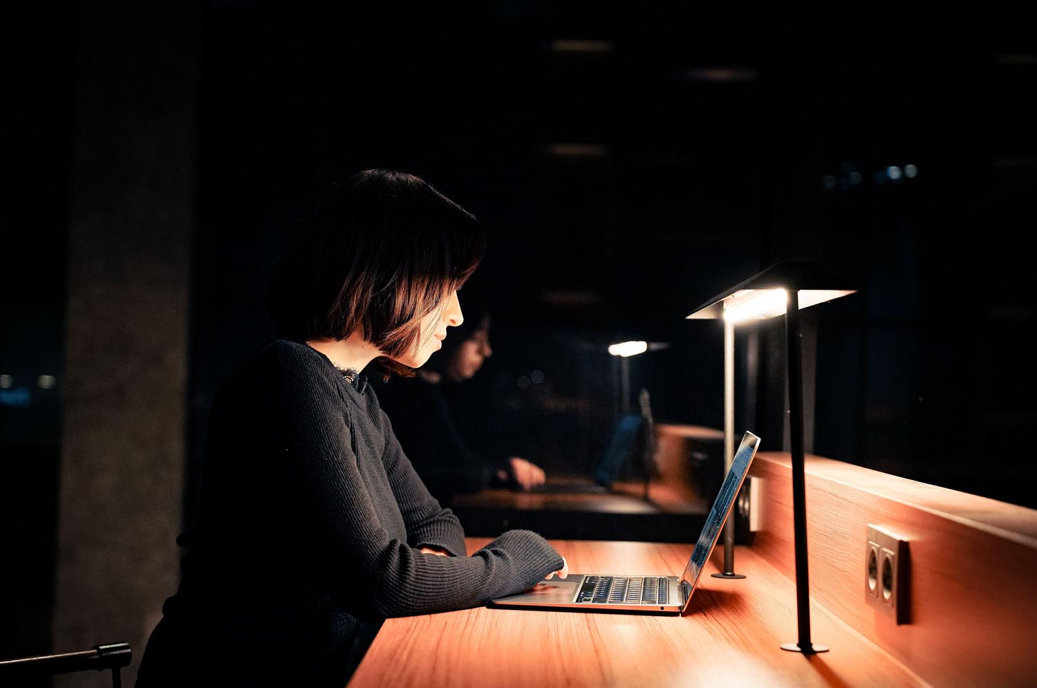 Mujer trabajando en una laptop en una habitación oscura, resaltando el contraste de la luz fría de la pantalla y la necesidad de la luz nocturna.