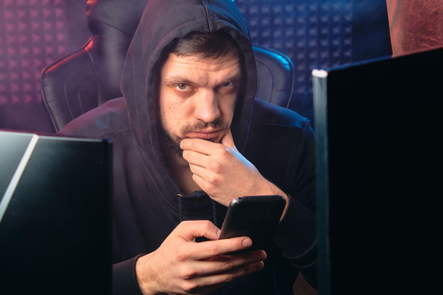 Hombre con capucha negra utilizando un smartphone en un entorno oscuro con pantallas digitales, simbolizando la amenaza de un ciberdelincuente.