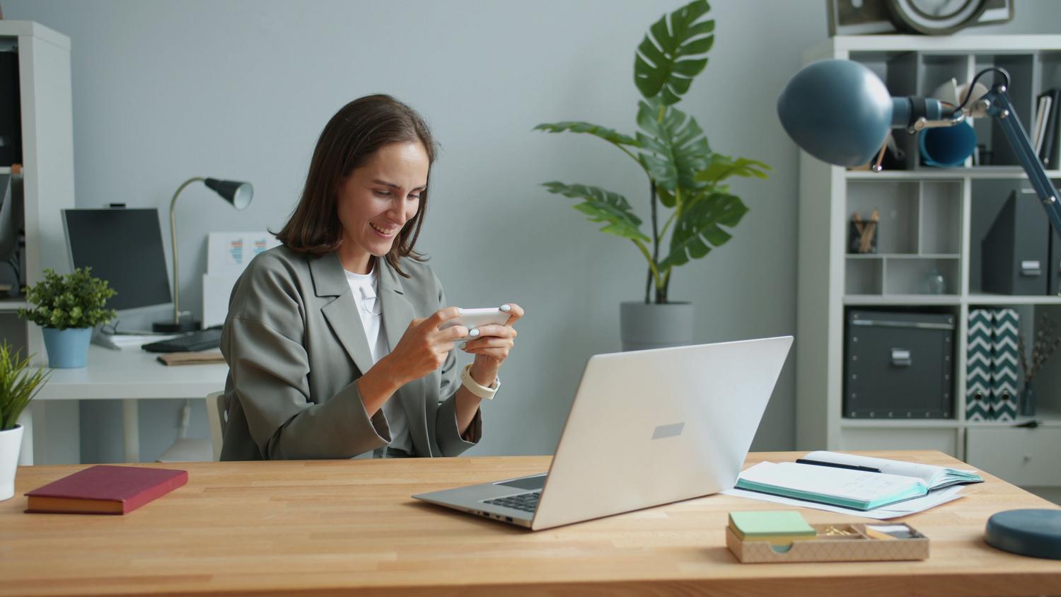 Mujer en su oficina en casa utilizando un smartphone y un ordenador portátil, reflejando la productividad del ecosistema digital sincronizado.