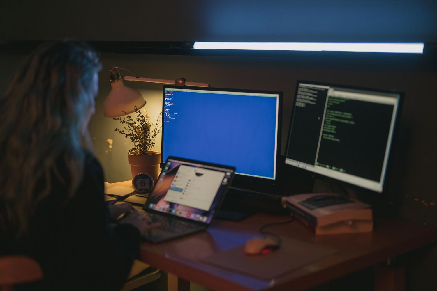Mujer utilizando múltiples monitores en una oficina en casa, con pantallas que muestran terminales de comandos y entornos técnicos.