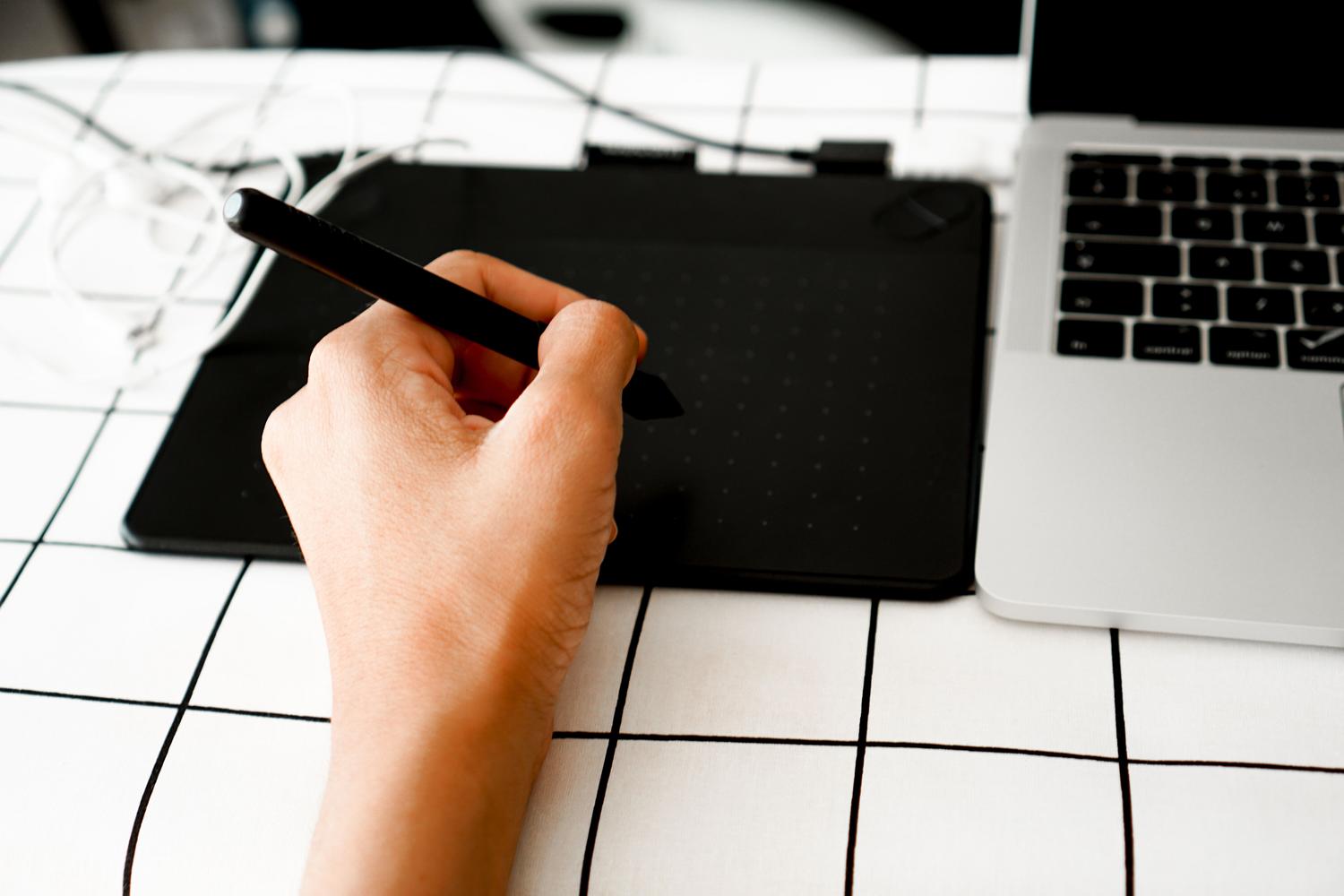 Mano utilizando un lápiz digital sobre una tableta gráfica en un escritorio blanco minimalista.