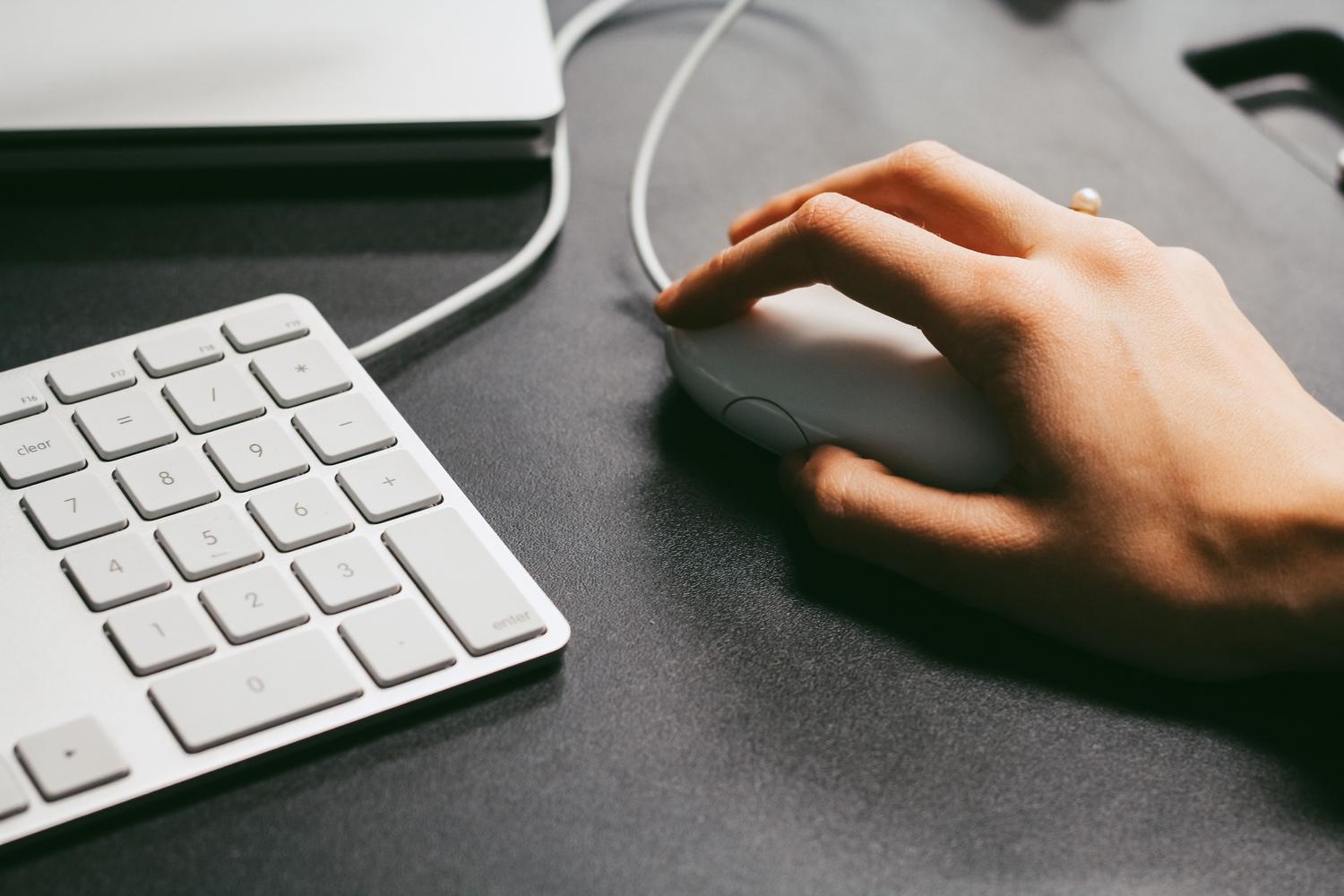 Mano operando un ratón de ordenador junto a un teclado blanco en un escritorio oscuro y minimalista. Mano operando un ratón de ordenador junto a un teclado blanco en un escritorio oscuro y minimalista.