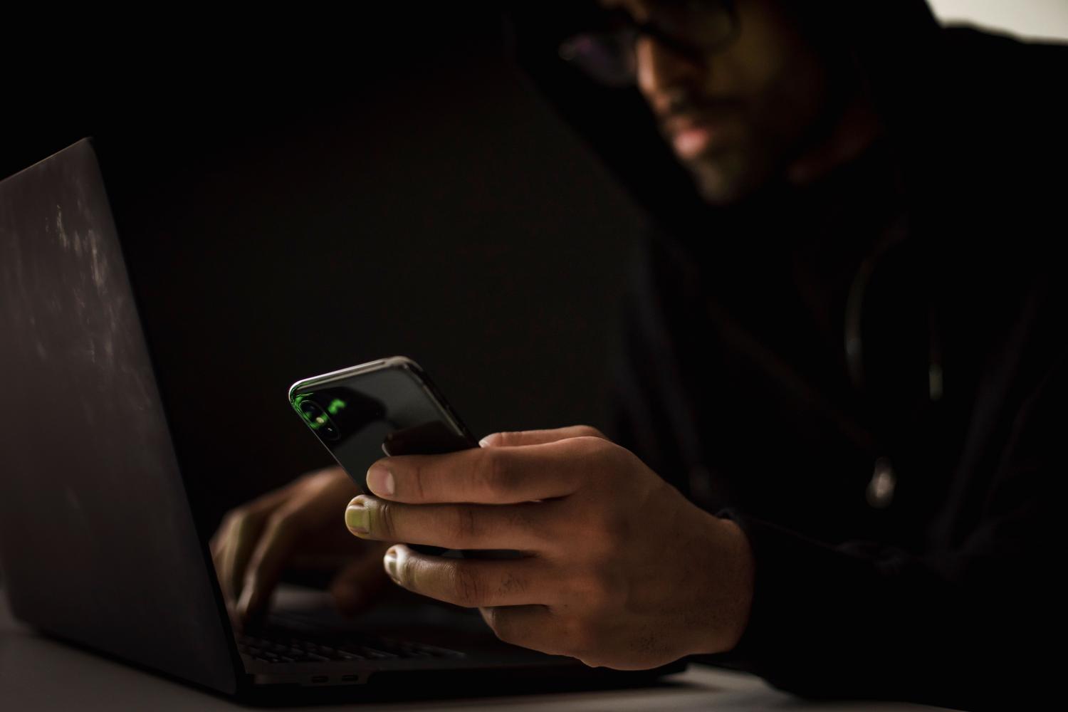 Persona experta en ciberseguridad utilizando un smartphone y un portátil en un entorno oscuro con reflejos de código verde en la pantalla.