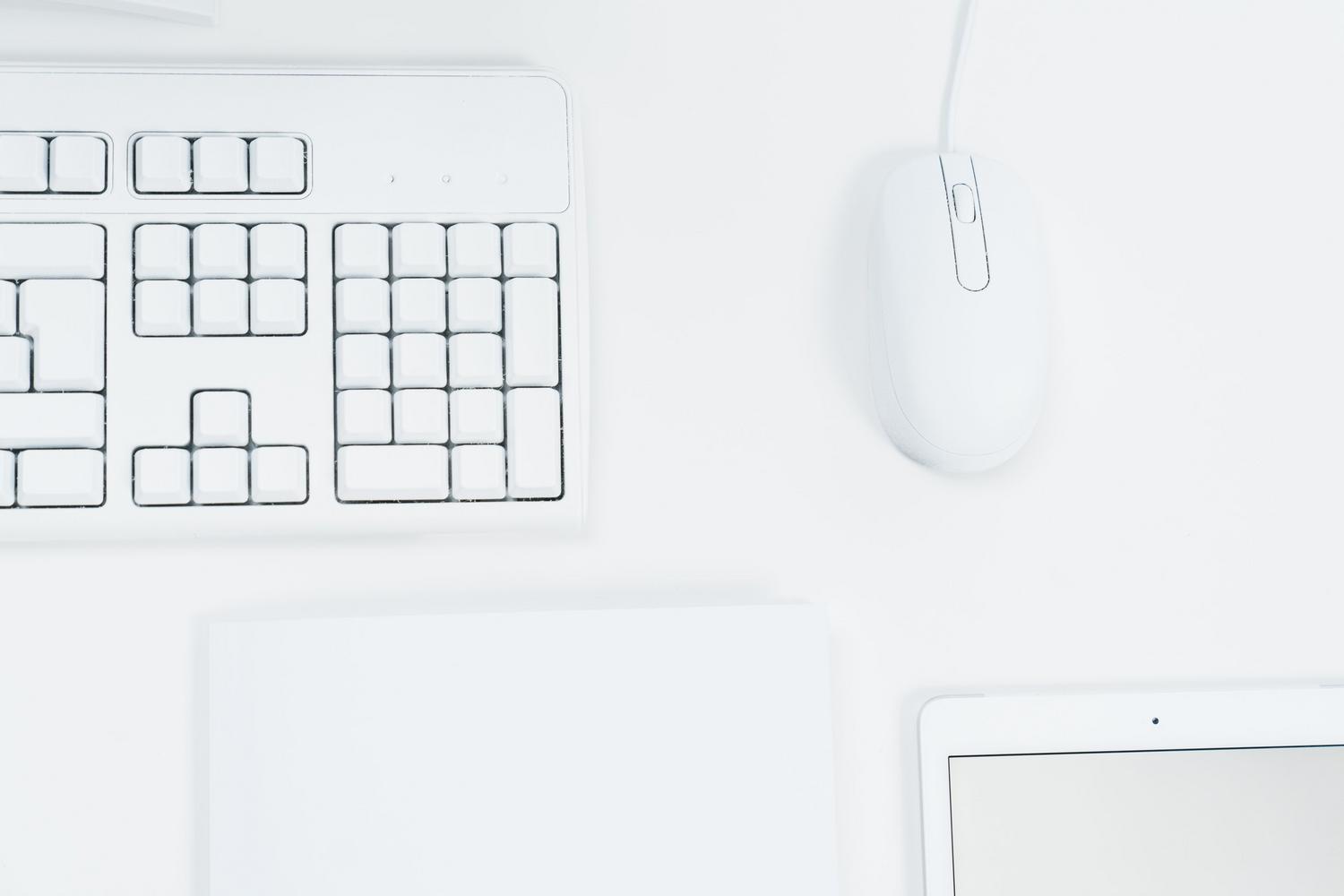 Vista cenital minimalista de un teclado, ratón y tablet sobre un escritorio blanco, simbolizando un espacio de trabajo eficiente.