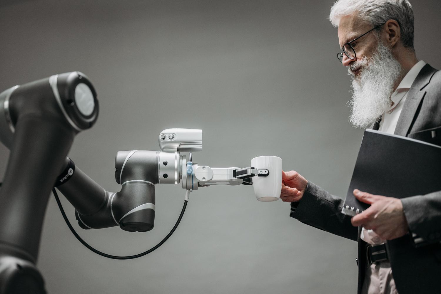 A robotic arm assists an elderly man by handing him a cup, symbolizing AI as human and inclusive support. A robotic arm assists an elderly man by handing him a cup, symbolizing AI as human and inclusive support.