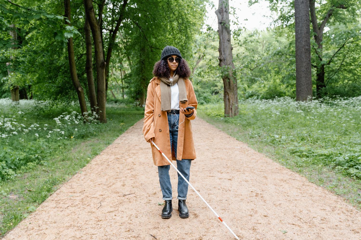 Mujer con bastón blanco y auriculares utilizando su smartphone para guiarse en un sendero natural.